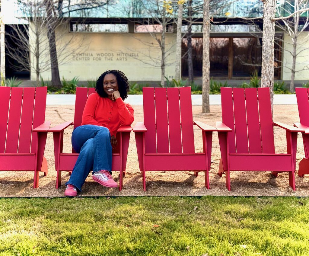 Black Women in red sweater, jeans, and pink shoes, sitting on a red beach chair outside on the lawn.